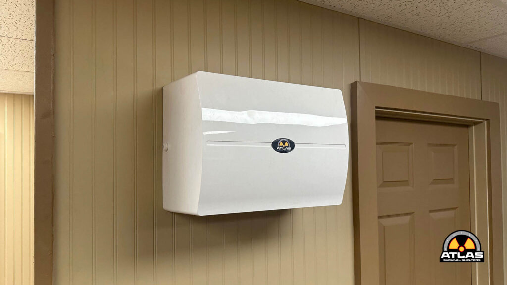 White wall-mounted Atlas shelter unit on a beige paneled wall beside a brown door; Atlas Survival Shelters logo visible on the front.