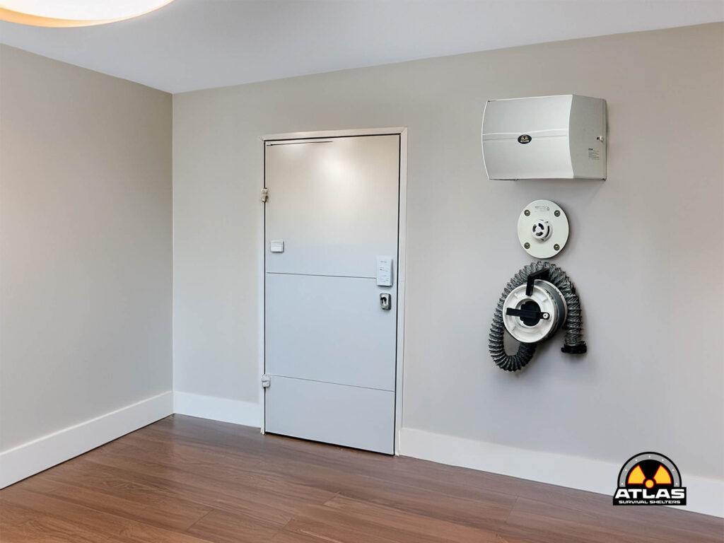 Corner of a utility room with a metal access door and wall-mounted equipment, including a coiled hose and Atlas Survival Shelters logo in the bottom corner.