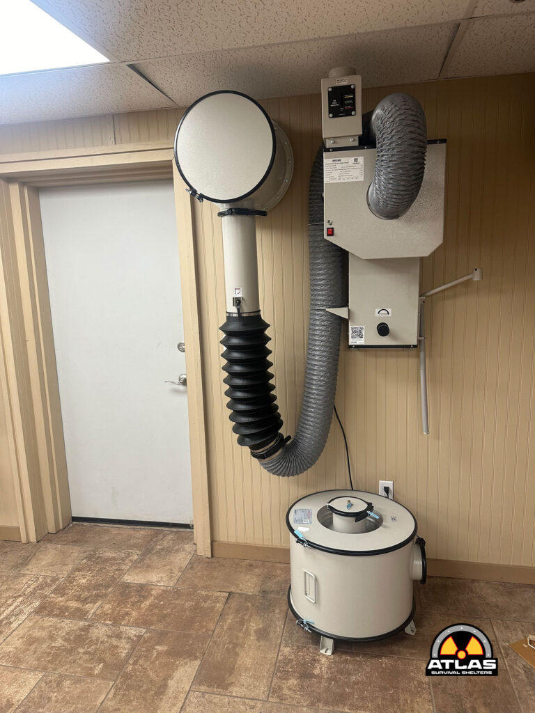 Industrial room with large ducting and a vertical vertical exhaust hose connected to a white cylindrical air handling unit; Atlas Survival Shelters logo visible in the bottom-right corner.