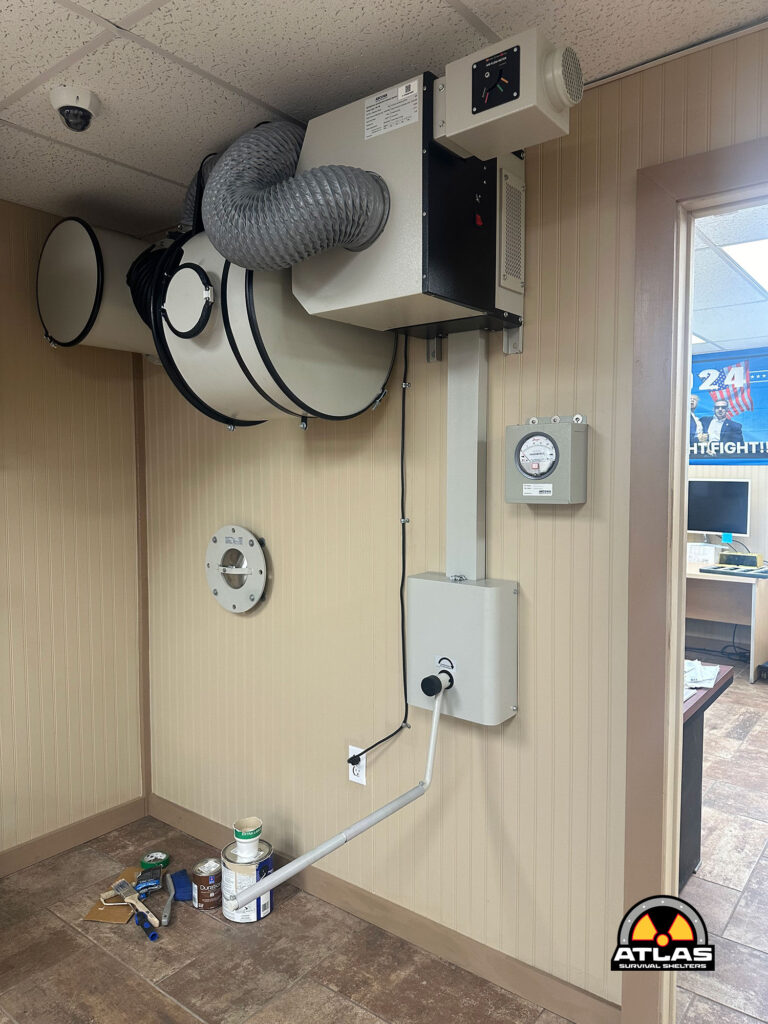 Industrial ventilation unit with flexible ducting and control box mounted on a beige paneled wall; tools and paint cans on the floor nearby.