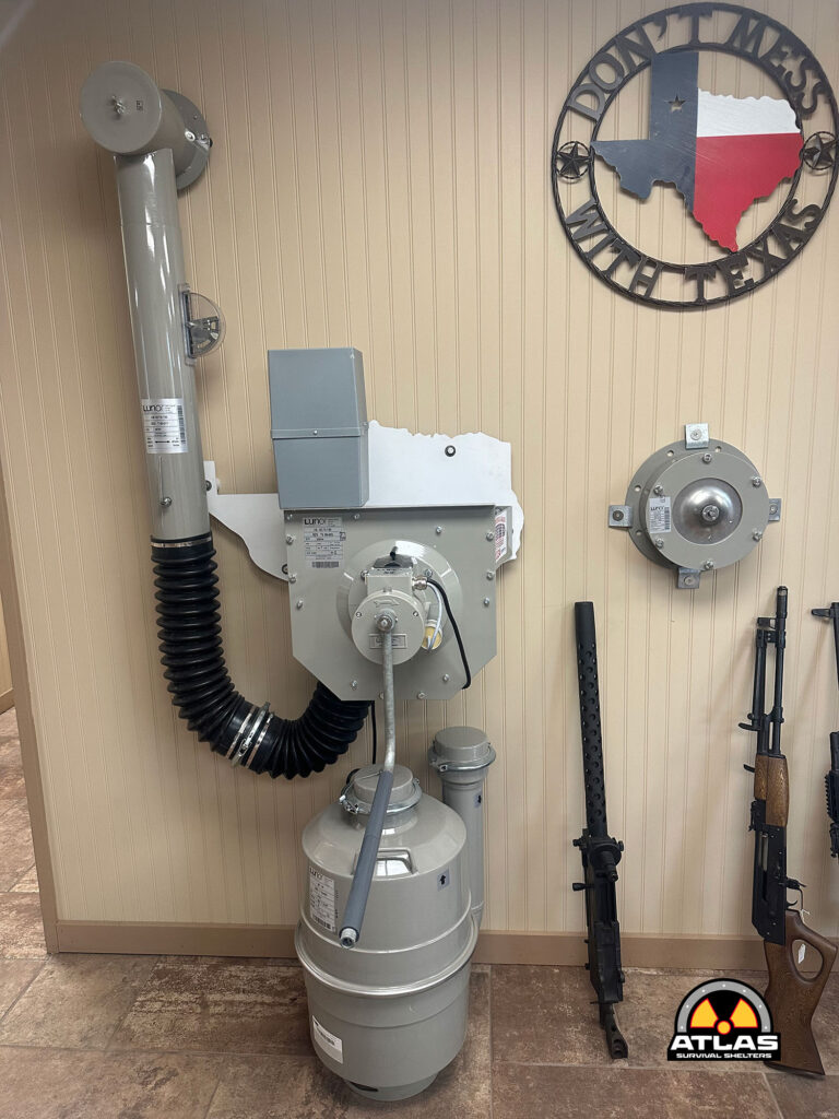 Wall-mounted display of shelter equipment: large gray vent duct with elbow, control box, propane tank, and rifles on stands, Atlas Survival Shelters logo in corner.