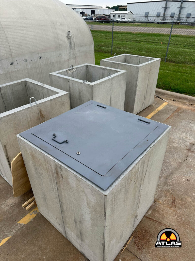 Four concrete square shelter modules with metal access lids in a paved outdoor lot, industrial buildings in the background.