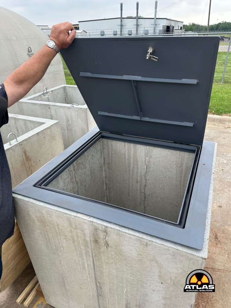 Person lifting a large metal hatch from a concrete underground shelter, with a lock visible on the door. An Atlas Survival Shelters logo appears at the bottom corner.