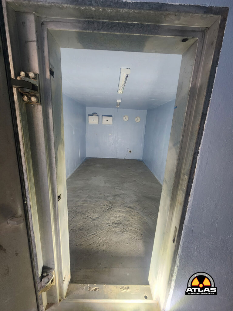 Interior of a blue-walled small shelter with a concrete floor, open metal hatch framing, and ceiling fluorescent lights illuminating the room.