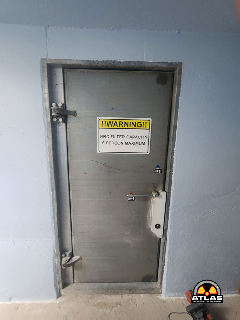 Metal door with a yellow warning sign: 'NBC FILTER CAPACITY 6 PERSON MAXIMUM'.