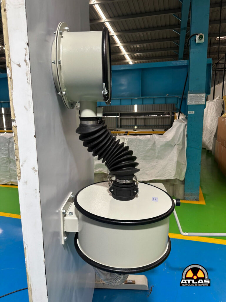 Industrial ventilation duct with a large circular flange and black accordion hose attached to a wall in a warehouse; Atlas Survival Shelters logo in the corner.