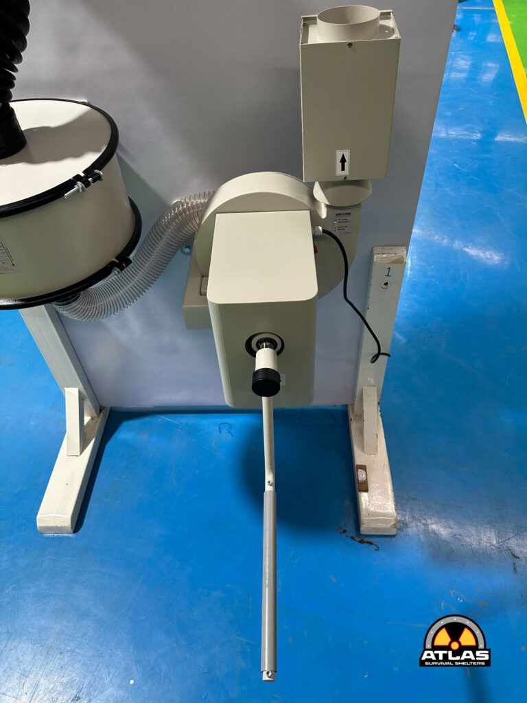 Industrial machine with a vertical metal rod and a gray control box, hose connections, on a blue workshop floor with an Atlas Survival Shelters logo in the corner.
