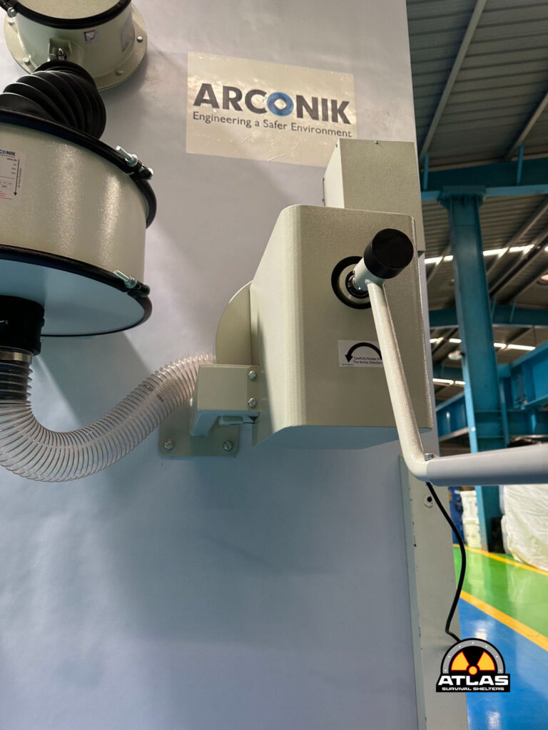 Industrial machine with a vertical lever, hoses, and a sign reading 'ARCONIK Engineering a Safer Environment' on a gray wall in a factory.