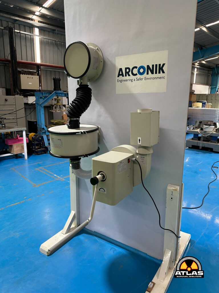 Industrial workshop scene with a beige, circular-hooded machine connected by a flexible hose to a vertical support, Atlas Survival Shelters logo in the corner on a blue floor.