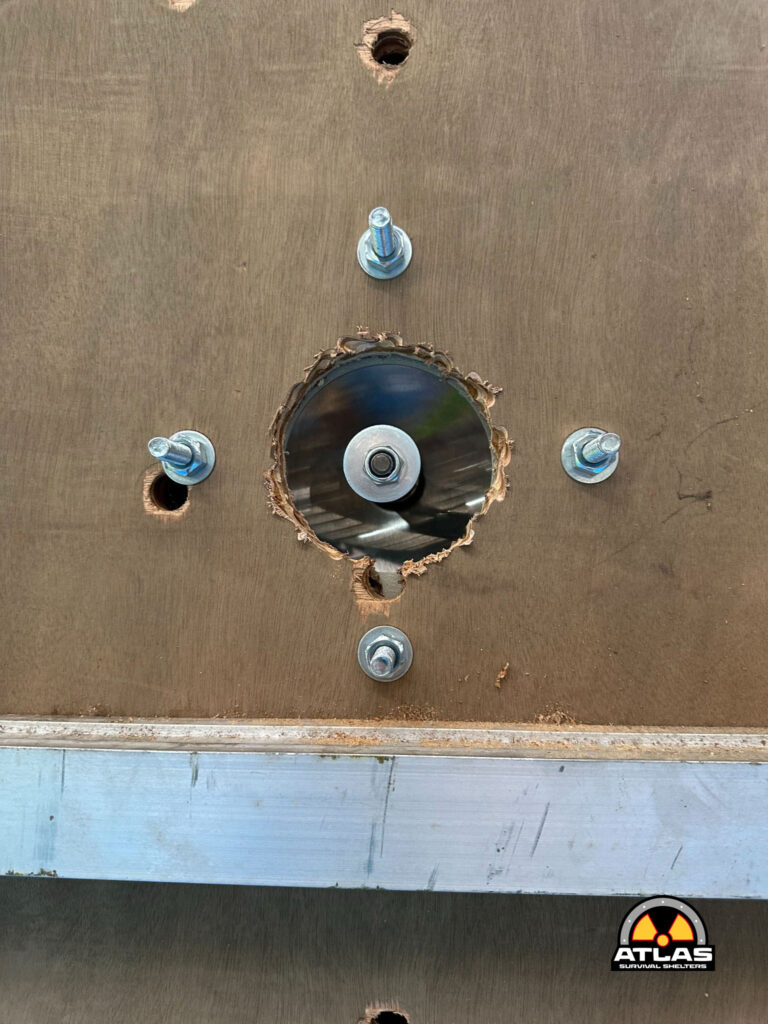 Plywood panel with a large circular hole and bolts around the opening; Atlas Survival Shelters logo in the bottom-right corner.