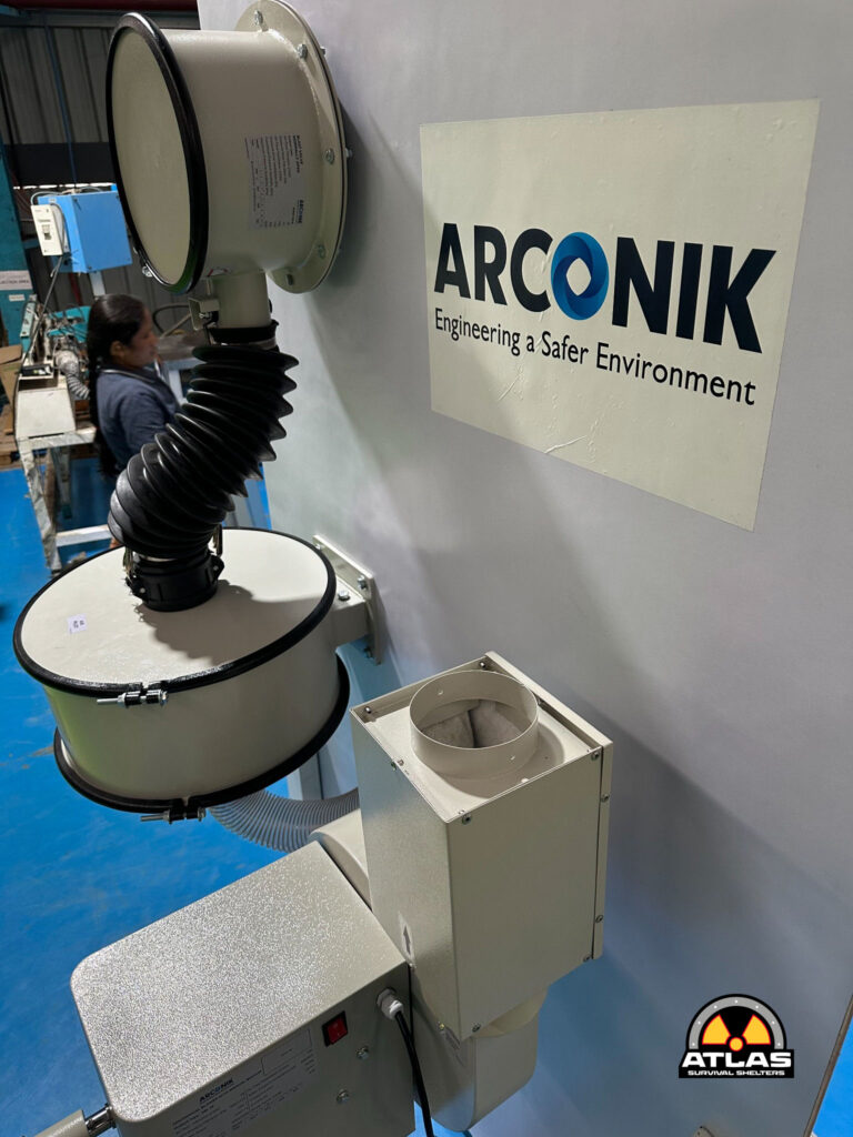 Industrial testing apparatus with a large beige cylindrical chamber and coiled black ducting, mounted against a wall with an ARCONIK sign nearby.