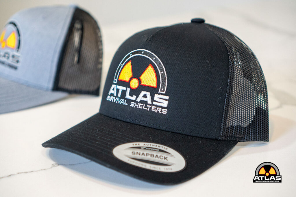 Black trucker hat featuring the Atlas Survival Shelters logo in white and yellow on the front, mesh back, resting on a white surface with a Snapback sticker visible on the bill.
