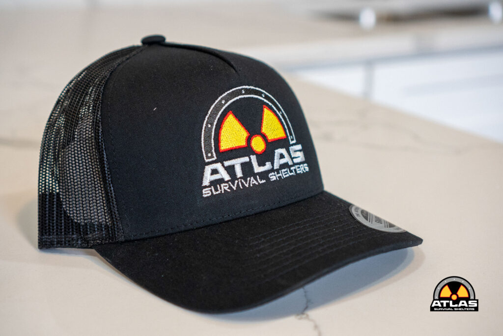 Black trucker cap with a front embroidered Atlas Survival Shelters logo (yellow radiation symbol) on a dark panel and mesh back.