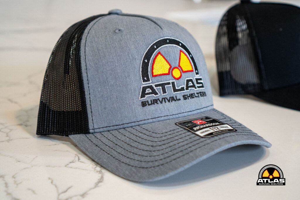Gray trucker hat with Atlas Survival Shelters logo embroidered on the front and a mesh back on a light countertop with a brim sticker visible.