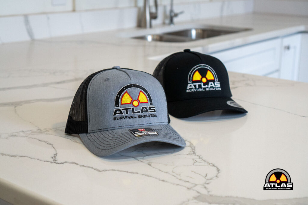 Two Atlas Survival Shelters trucker caps on a marble kitchen island, one gray in front and one black behind, logos visible on both hats.