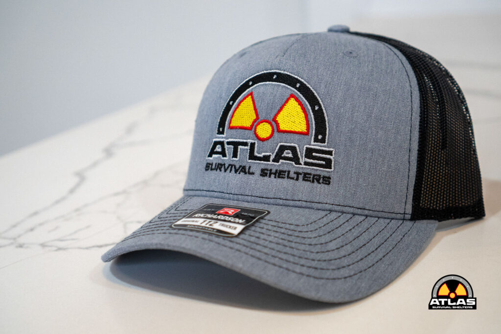 Blue trucker cap with Atlas Survival Shelters logo featuring a yellow radiation symbol on the front panel, mesh back, and sticker on the brim.