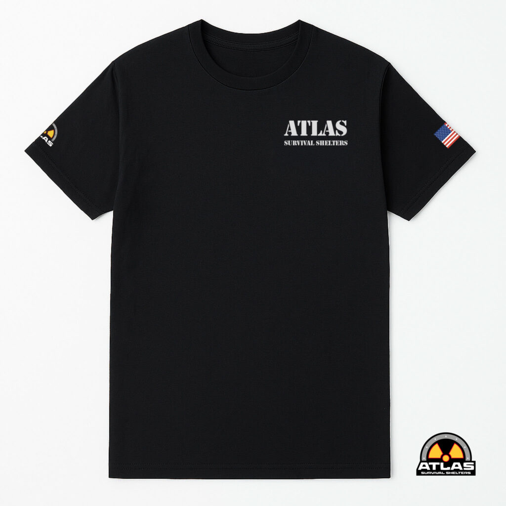 Black T-shirt showing the Atlas Survival Shelters logo on the chest, with an American flag patch on the right sleeve and a radium-style patch at the bottom-right corner.