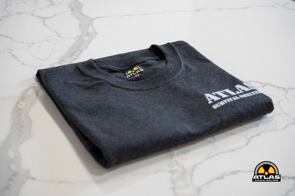 Folded charcoal T-shirt on a marble surface with an Atlas Survival Shelters logo on the chest area and a round patch at the bottom corner of the image.