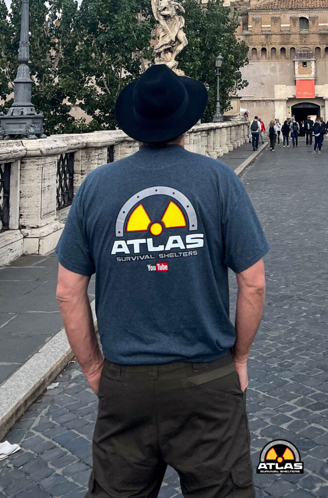 Back view of a man wearing a dark hat and a blue Atlas Survival Shelters T-shirt on a stone bridge; logo on shirt and a smaller patch logo on the ground nearby.