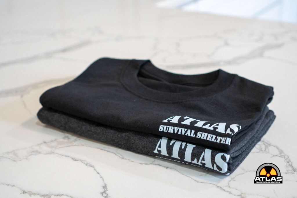Stack of folded black T-shirts on a marble surface, front shirt reads 'ATLAS SURVIVAL SHELTERS' with a round logo in the bottom-right corner of the image.
