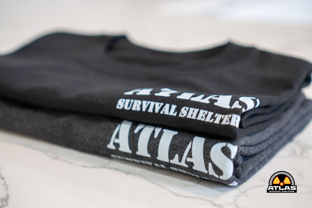Close-up of folded black shirts with bold white text reading 'SURVIVAL SHELTERS' and the Atlas logo visible in the bottom right corner.