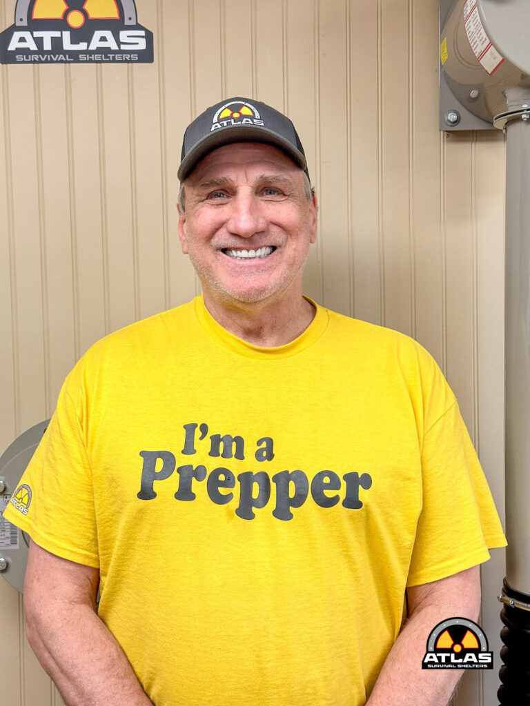 Smiling man in a yellow t‑shirt that reads I'm a Prepper, standing in front of a beige wall with Atlas Survival Shelters logos on hat and sign nearby