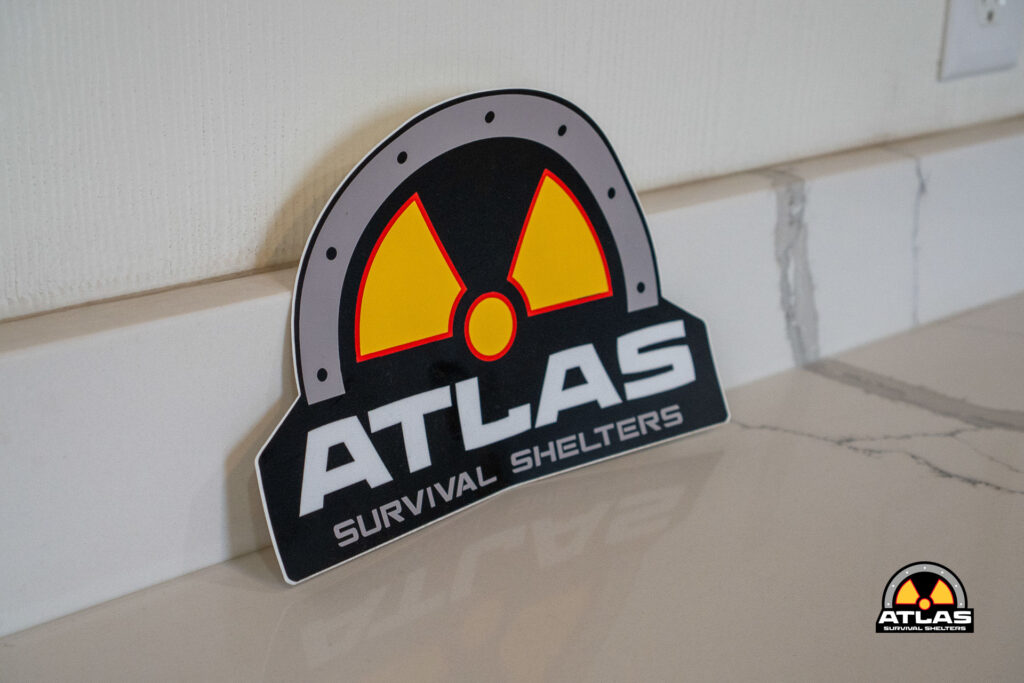 Tilted sign reading 'ATLAS Survival Shelters' with a yellow and red radiation symbol, placed on a light-colored floor beside a white wall.