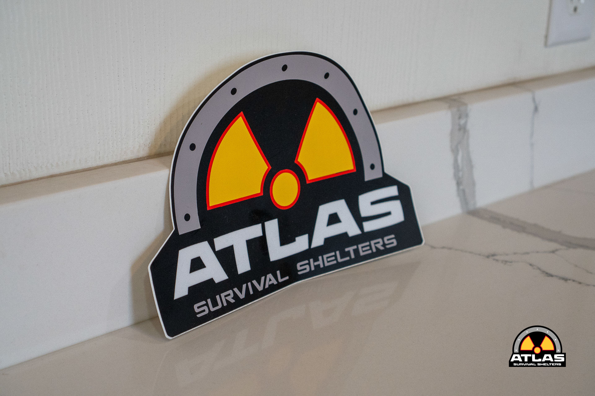 Tilted sign reading 'ATLAS Survival Shelters' with a yellow and red radiation symbol, placed on a light-colored floor beside a white wall.