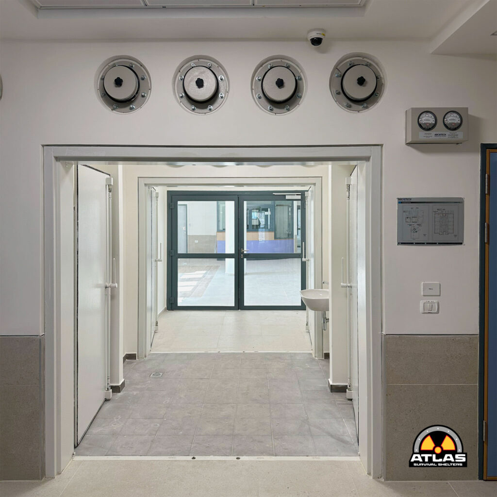 Interior corridor with four circular wall access ports above a doorway; security camera overhead and an Atlas Survival Shelters panel on the right.