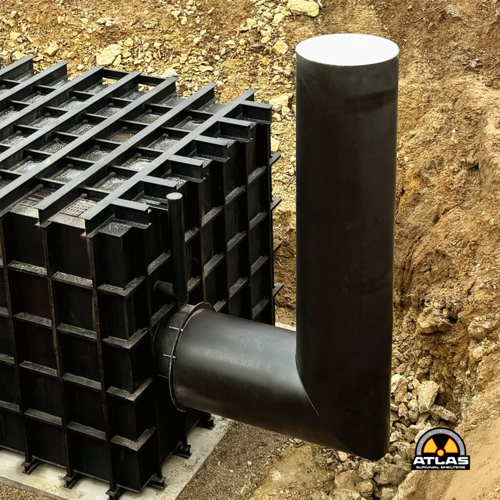 Stacks of black metal formwork panels arranged in a grid beside a large black cylindrical pipe on a dirt construction site, with the Atlas Survival Shelters logo in the bottom-right corner.