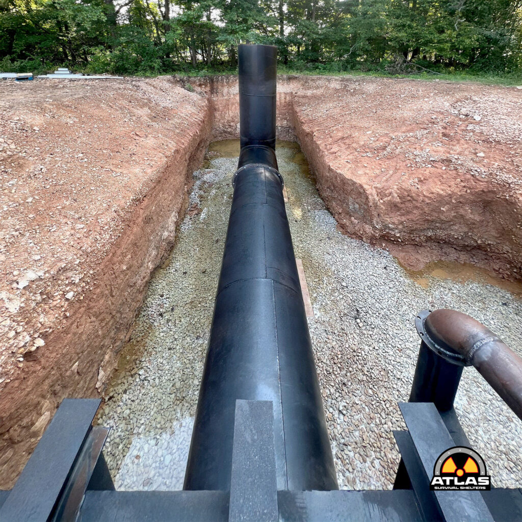 Underground trench with a large black pipe laid across, dirt walls on both sides; Atlas Survival Shelters logo bottom-right.