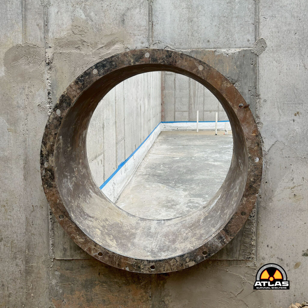 Rusty circular metal ring embedded in a rough concrete wall, with the Atlas Survival Shelters logo in the bottom-right corner.