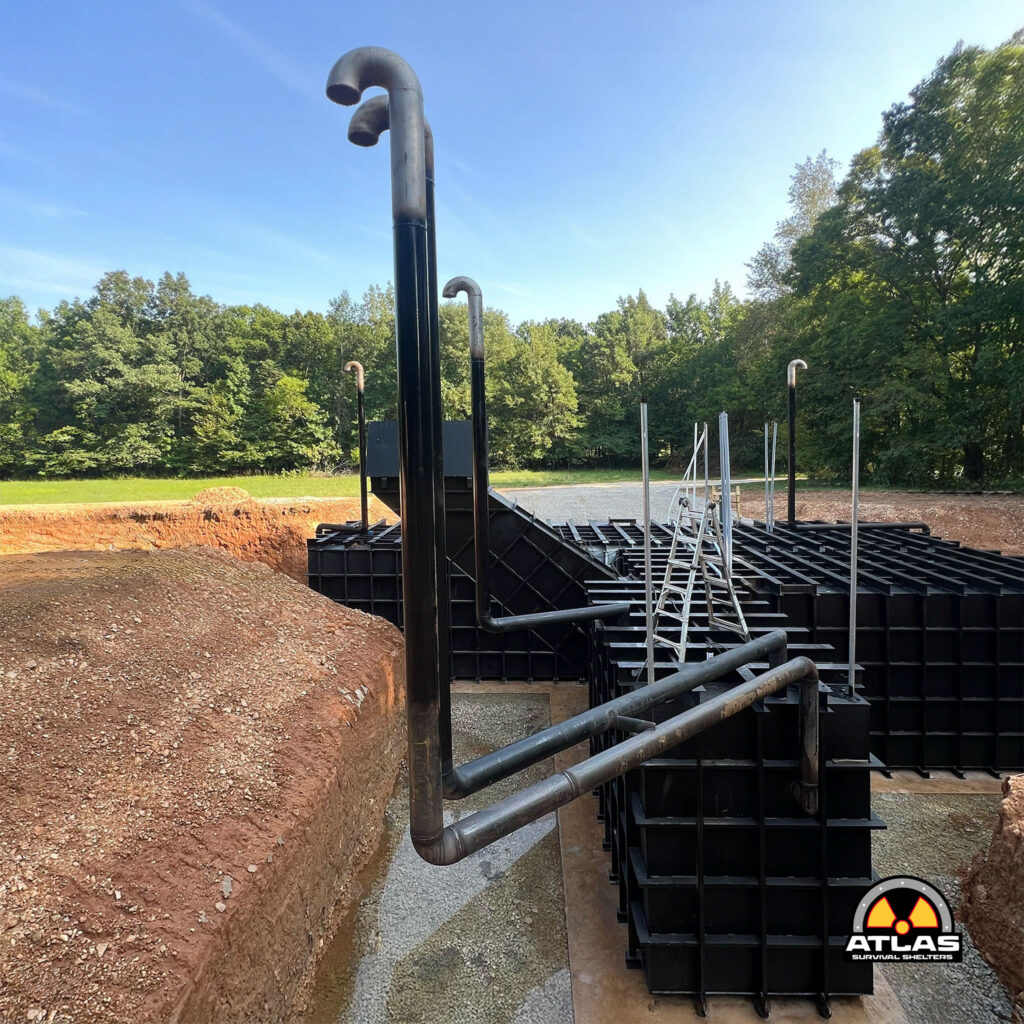 Industrial construction site with black modular containers and tall curved pipes, Atlas Survival Shelters logo visible in bottom right.