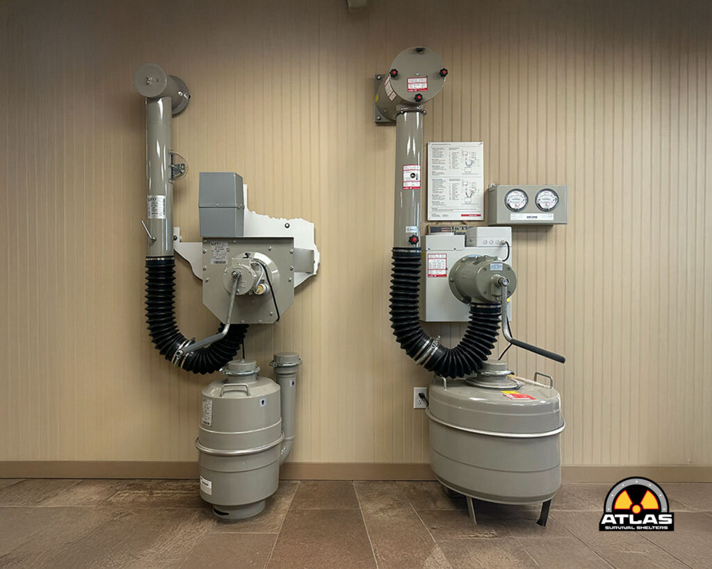 Two large gray gas cylinders connected to wall-mounted equipment with black corrugated hoses in a beige utility room; gauges and controls nearby (Atlas logo in corner).
