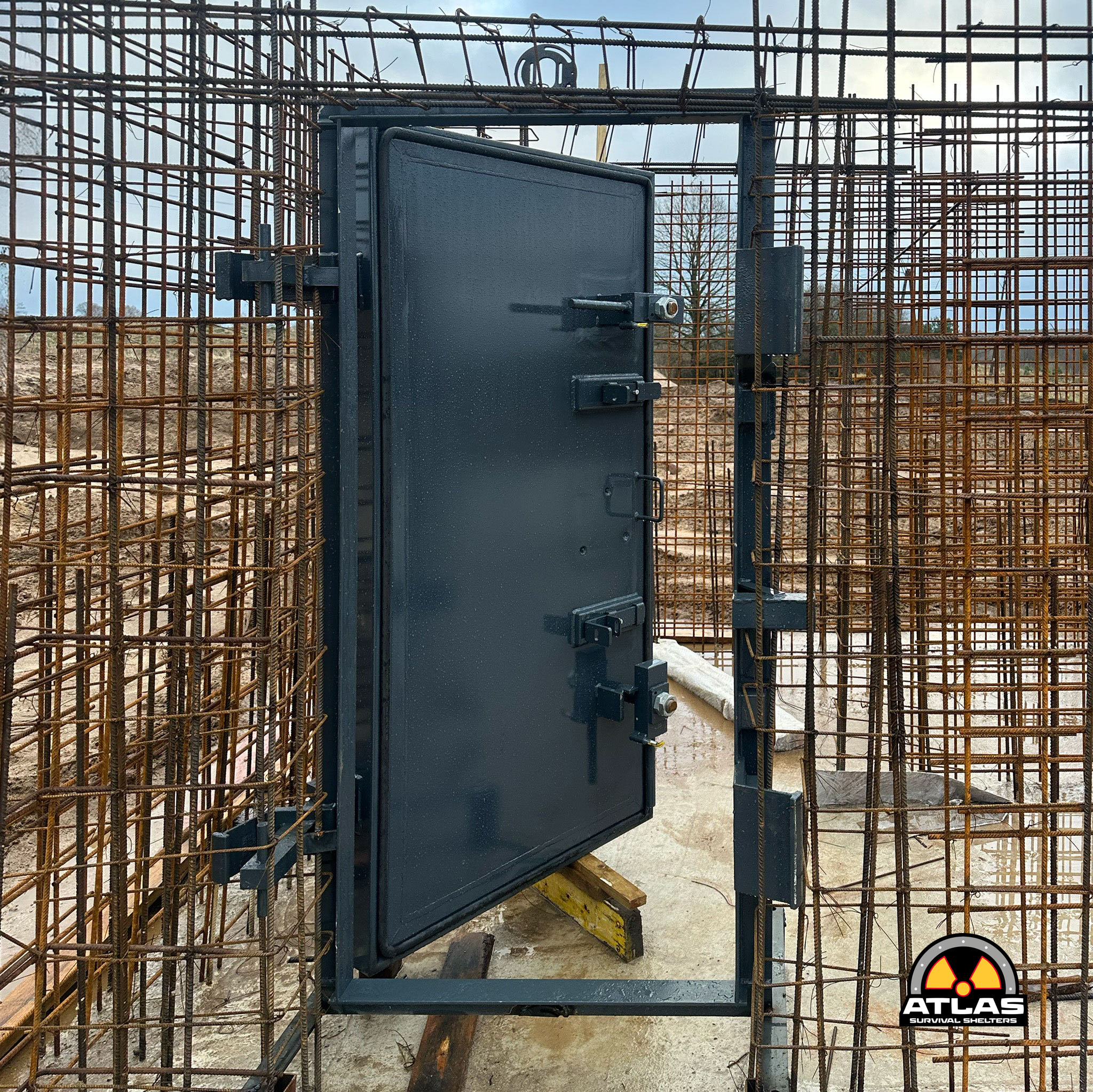 Industrial construction scene with a dark gray metal shelter door surrounded by rusted rebar mesh; safe room in progress under concrete slab footing.