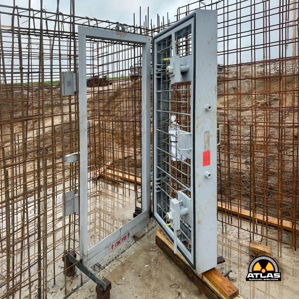 Steel-framed shelter being installed on a rebar-filled construction site; concrete foundation and ATLAS Survival Shelters logo visible in the corner.