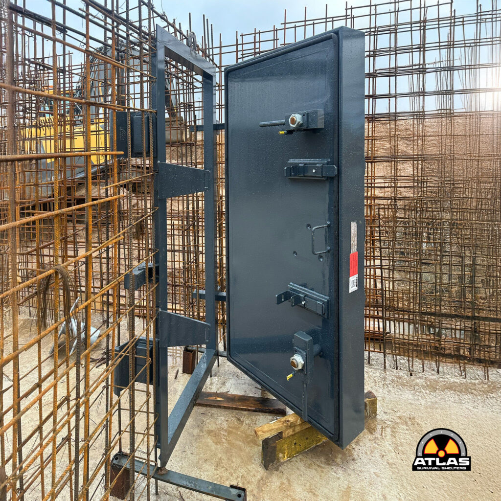 Construction site with exposed steel reinforcement bars and a gray metal shelter panel in the foreground, Atlas Survival Shelters logo visible at the bottom right.