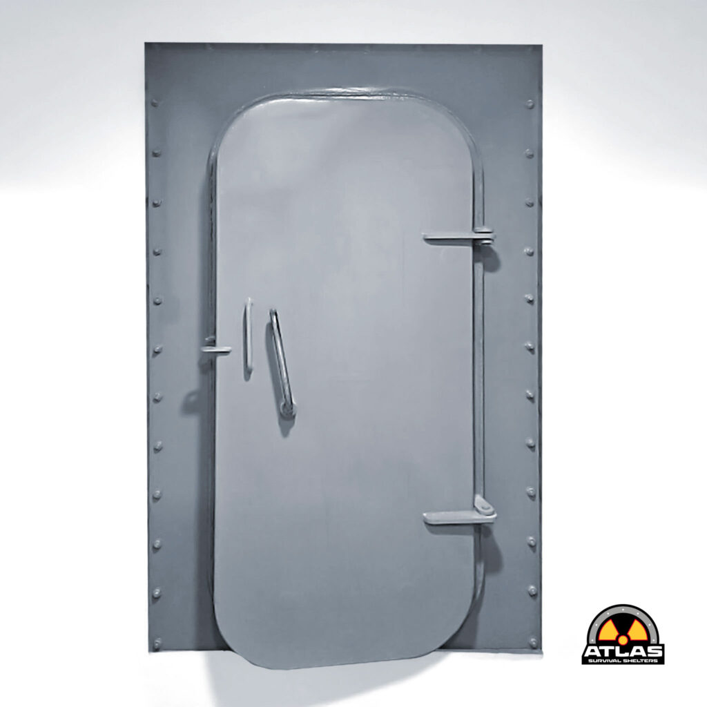 Grey metal blast shelter door with a rounded inner hatch and vertical handle; Atlas Survival Shelters logo bottom-right corner.