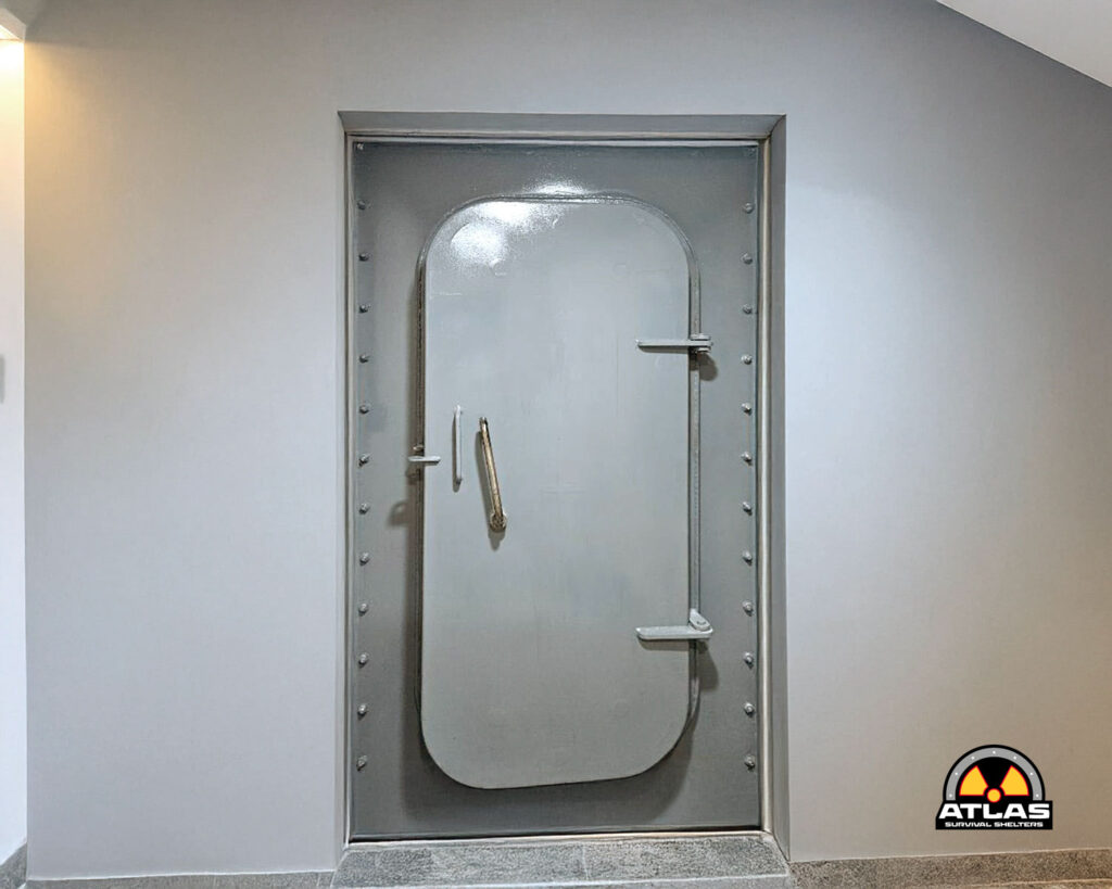 Heavy steel bunker door with a rounded top inset in a gray wall, featuring a vertical handle and riveted frame bolts on both sides.