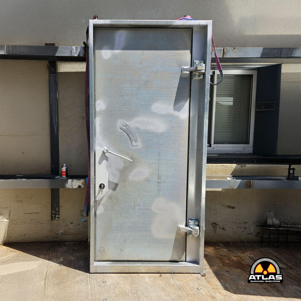 Metal utility door or cabinet mounted outside against a stucco wall, with a padlock and cables visible, plus an Atlas Survival Shelters logo in the bottom-right corner.