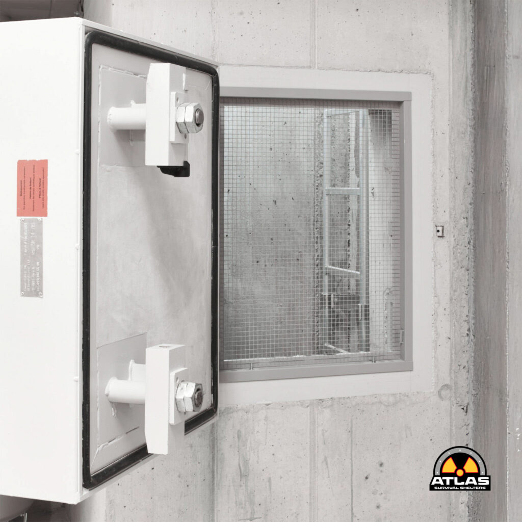 White industrial vault door with a prominent locking bolt mounted on a concrete wall, next to a small windowed section; Atlas Survival Shelters logo in the bottom-right corner.