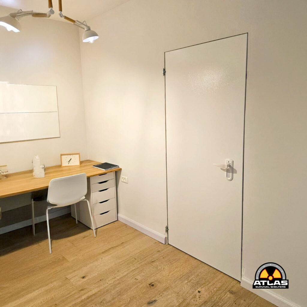 Office workspace with a wooden desk, white chair, and a row of white cabinets along the wall; Atlas Survival Shelters logo in the bottom-right corner.