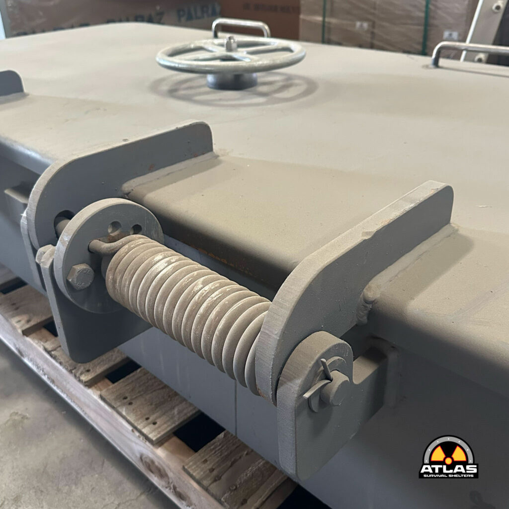 Close-up of a gray industrial roller assembly with round gears and a coiled cable, part of a metal shelter frame; Atlas Survival Shelters logo visible in the corner.