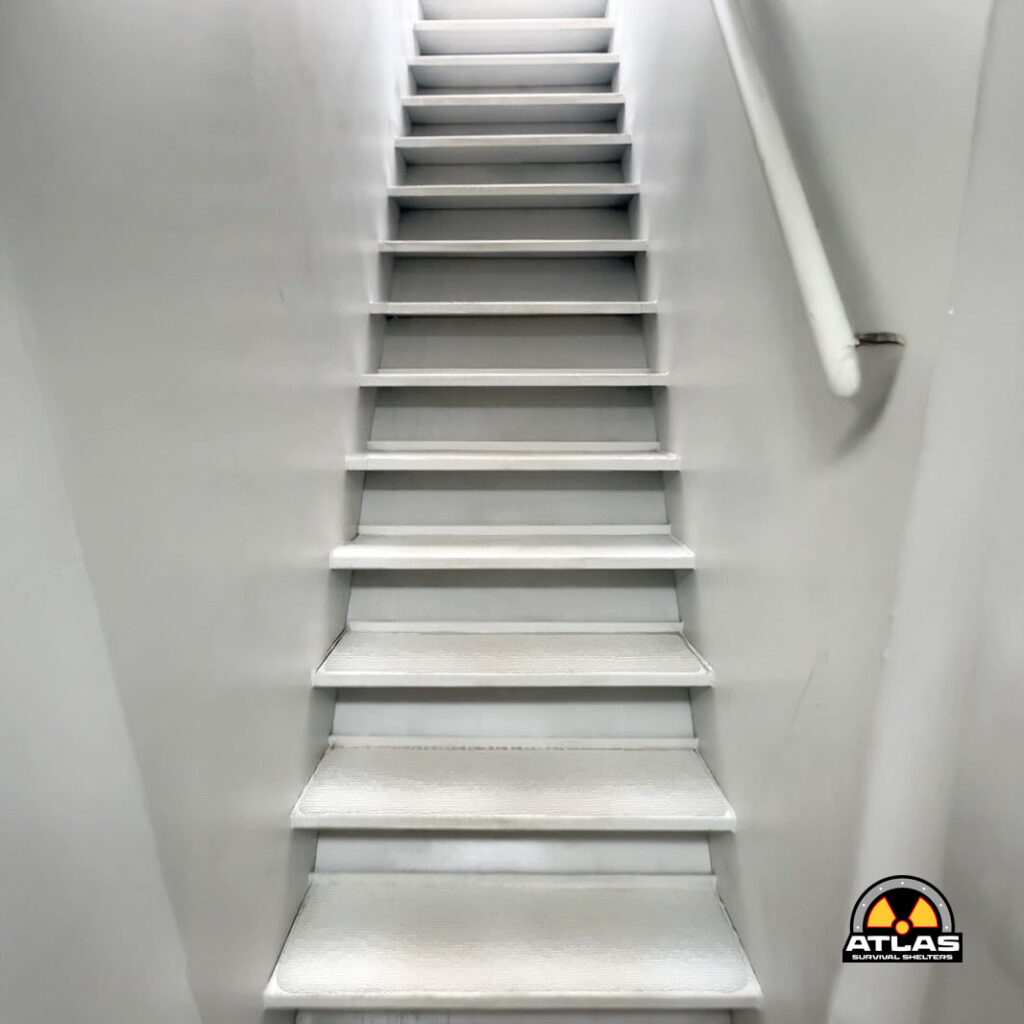 White metal staircase ascending in a narrow stairwell with a white handrail on the right; Atlas Survival Shelters logo visible in the bottom-right corner.